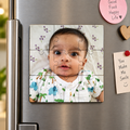 A square jigsaw puzzle magnet featuring a baby's portrait, shown attached to a stainless steel refrigerator door alongside other kitchen magnets and sticky notes.